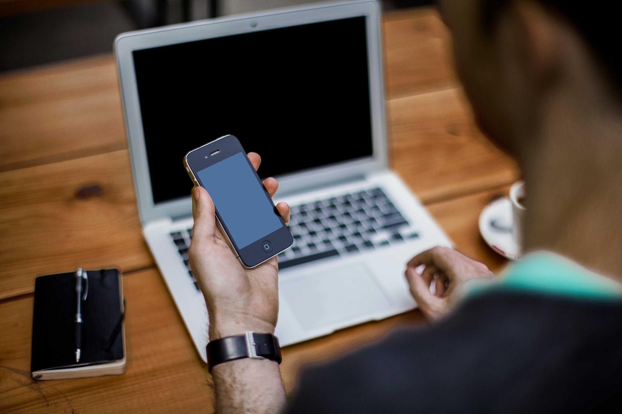Person checking a phone in front of a laptop at a wooden desk